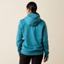 Back view of a person wearing a light teal hoodie with black pants against a beige background.