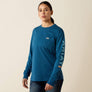 Person wearing a dark blue long sleeve shirt that has Ariat going down the arm in a light blue color and has a pocket on the chest area against a cream colored background.