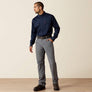 Man wearing a navy blue button up shirt and grey pants against a tan background