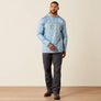 Man wearing a blue camo hoodie with Ariat in yellow on front with dark jeans on a beige background