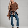 Person wearing a brown sweater that has buttons going down 1/4 of the front of the sweater and blue jean pants and also has a brown and tan flannel tied around their waste against a light grey background.