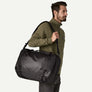 Person in an olive green fleece and grey pants carrying a black duffel bag over his shoulder against a white background.