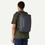 A Person in a green shirt and blue jeans wearing a grey backpack that says Patagonia on the front against a white background.