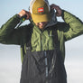 Person wearing a green jacket, black overalls, and a yellow cap with a logo against a clear sky.