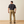 Backs view of a man wearing a dark grey shirt, khaki pants with boots against a light grey background and wood floor.