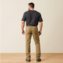 Backs view of a man wearing a dark grey shirt, khaki pants with boots against a light grey background and wood floor. 