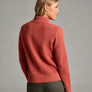 Back view of a person wearing a burnt orange sweater and green pants against a grey background.