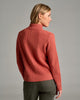 Back view of a person wearing a burnt orange sweater and green pants against a grey background.