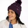 Person wearing a dark purple winter hat with a pom pom on the top in the same color and a light purple tag on the flipped up part of that hat.