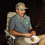 Person sitting in a lawn chair wearing a grey short sleeve button up shirt eating BBQ food at night