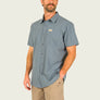Person wearing a grey button up short sleeve button up shirt with Marsh Wear patch on right side with tan pants on a blank background 