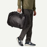 Bottom half of a person wearing an olive green sweatshirt, dark grey pants and grey and white shoes carrying a black duffel bag with his hand against a white background.
