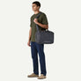 A person in a green shirt and blue jeans wearing an over the shoulder bag that is grey and have Patagonia on the front of it against a white background.