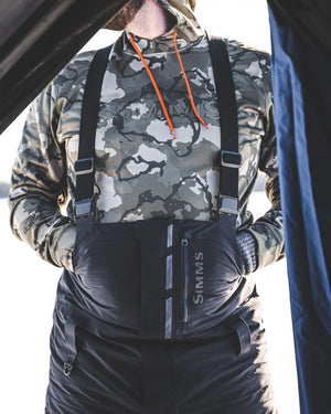 Person wearing camouflage jacket and black suspenders with 'Simms' branding.