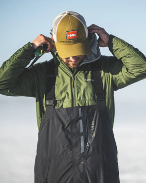 Person wearing a green jacket, black overalls, and a yellow cap with a logo against a clear sky.