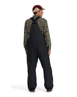 Person wearing a plaid shirt and black Simms Challenger Insulated bibs on a white background