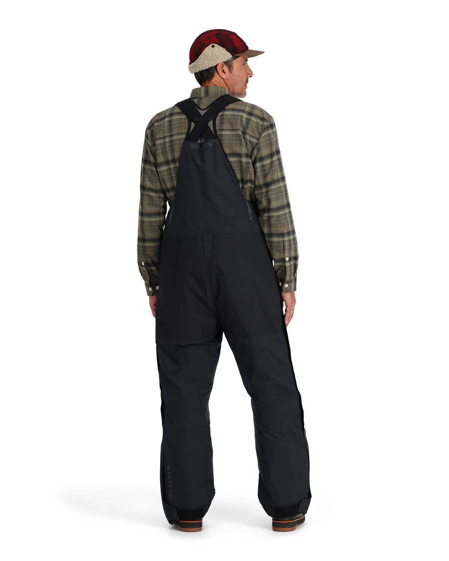 Person wearing a plaid shirt and black Simms Challenger Insulated bibs on a white background