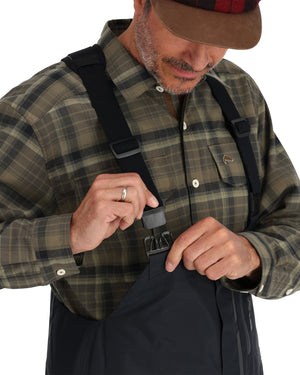 Man adjusting a black strap on Simms Challenger Insulated Bibs, wearing a plaid shirt and cap.
