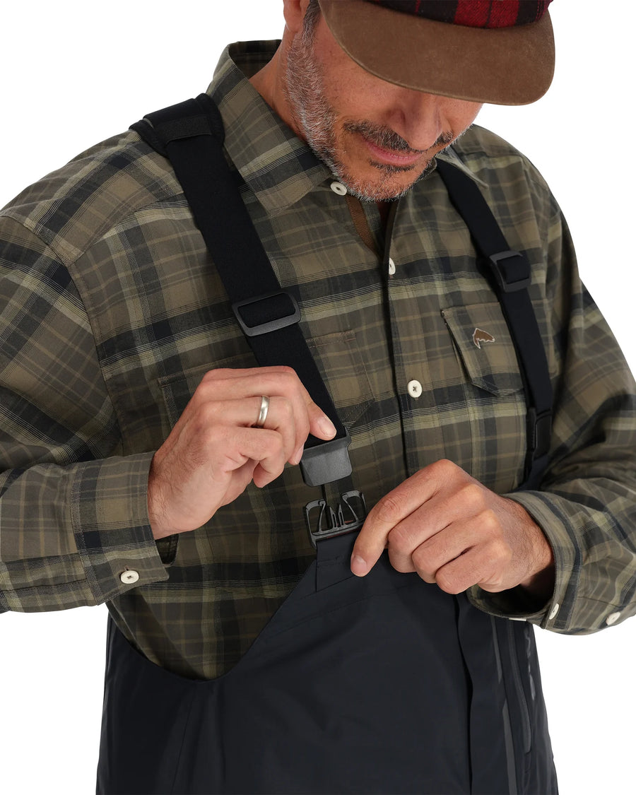 Man adjusting a black strap on Simms Challenger Insulated Bibs, wearing a plaid shirt and cap.