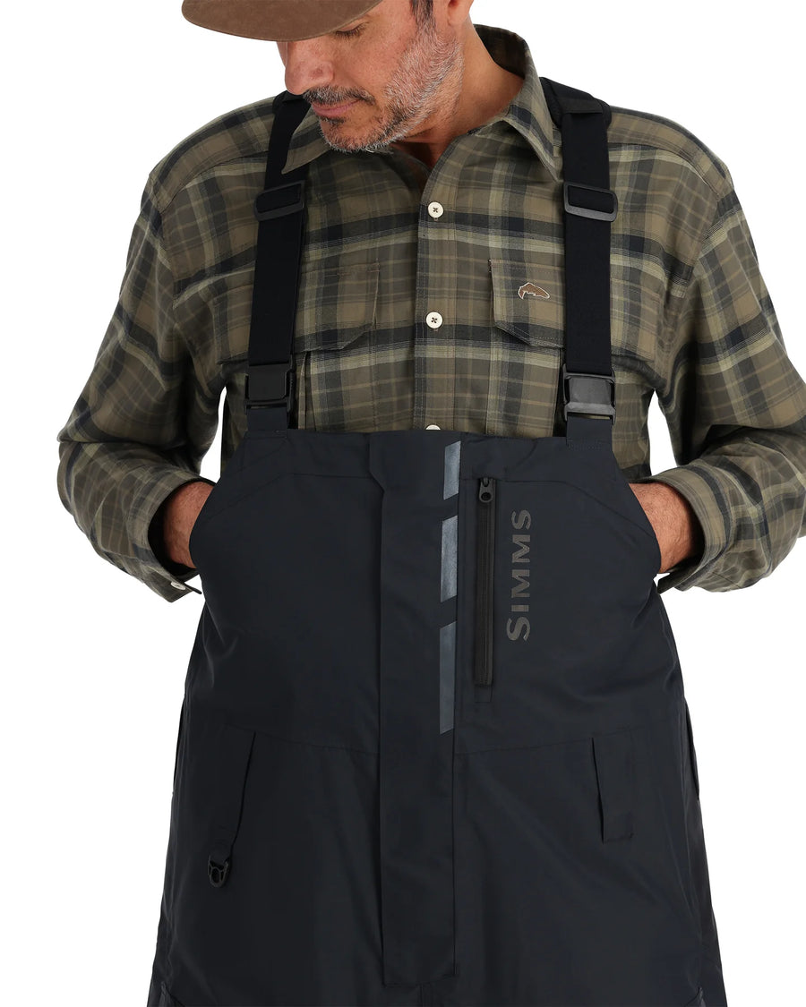 Man wearing a plaid shirt and black Simms fishing bib overalls on a white background
