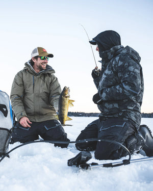 Two men ice fishing on a frozen lake with one holding a fish wearing Simms Challenger Jackets