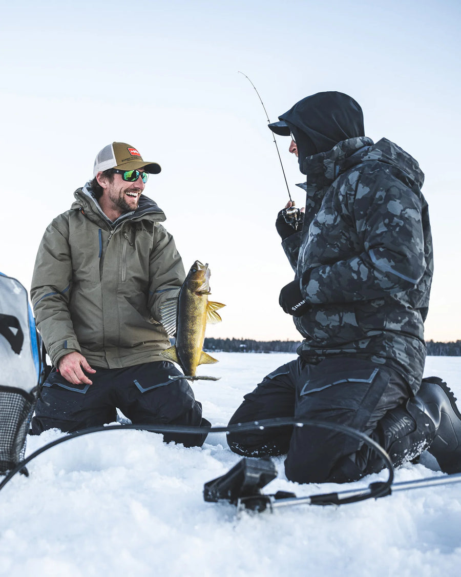 Two men ice fishing on a frozen lake with one holding a fish wearing Simms Challenger Jackets
