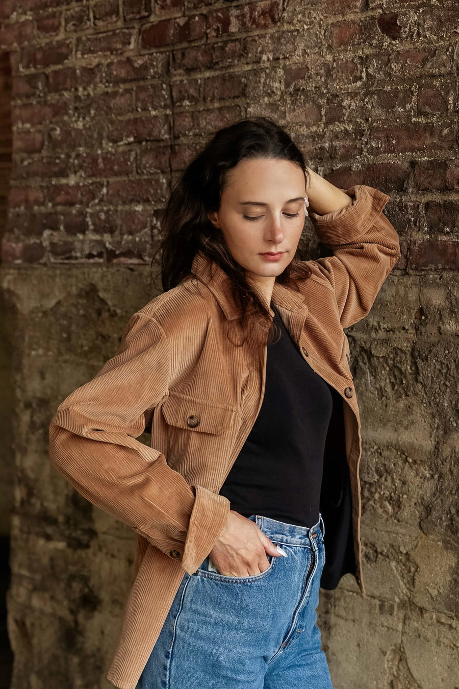 Woman wearing a brown jacket and blue jeans against a brick wall.